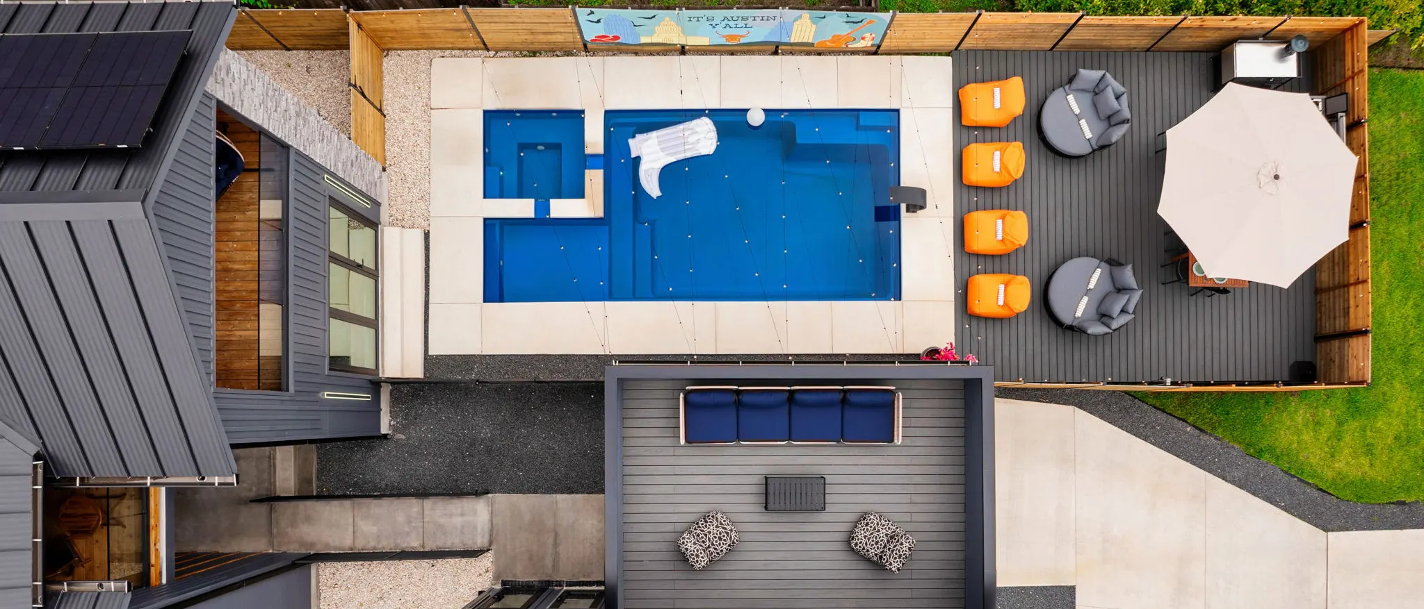 Aerial view of the backyard: the pool, the extended deck with orange loungers and daybeds, and the balcony off the bunk room with a navy sofa