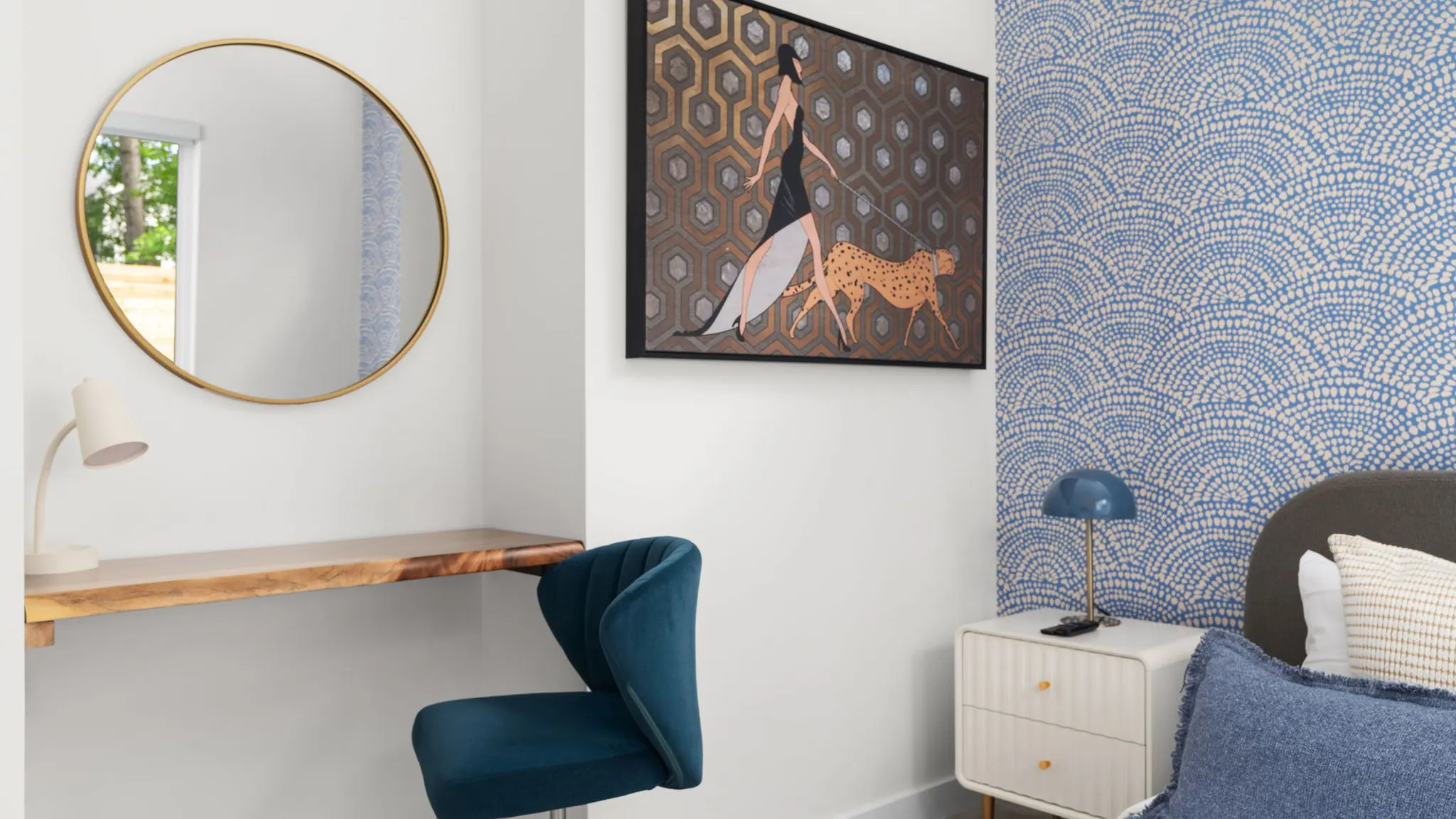 A bedroom desk with a teal velvet chair and a round gold mirror, beneath a painting of a woman walking a cheetah, next to blue scalloped wallpaper