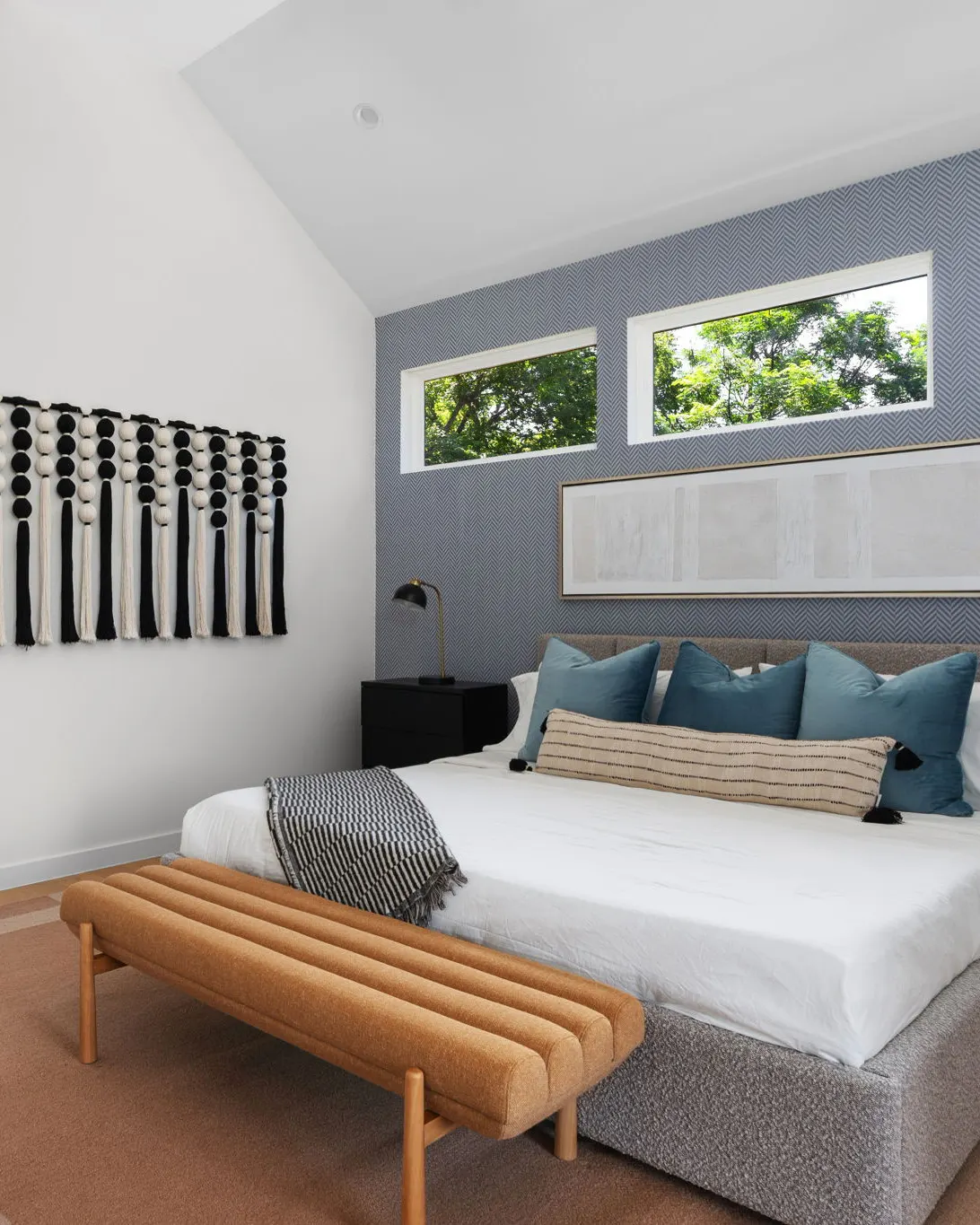 A bedroom with a navy herringbone accent wall, high windows onto the trees, teal pillows and a camel bench at the foot of the bed