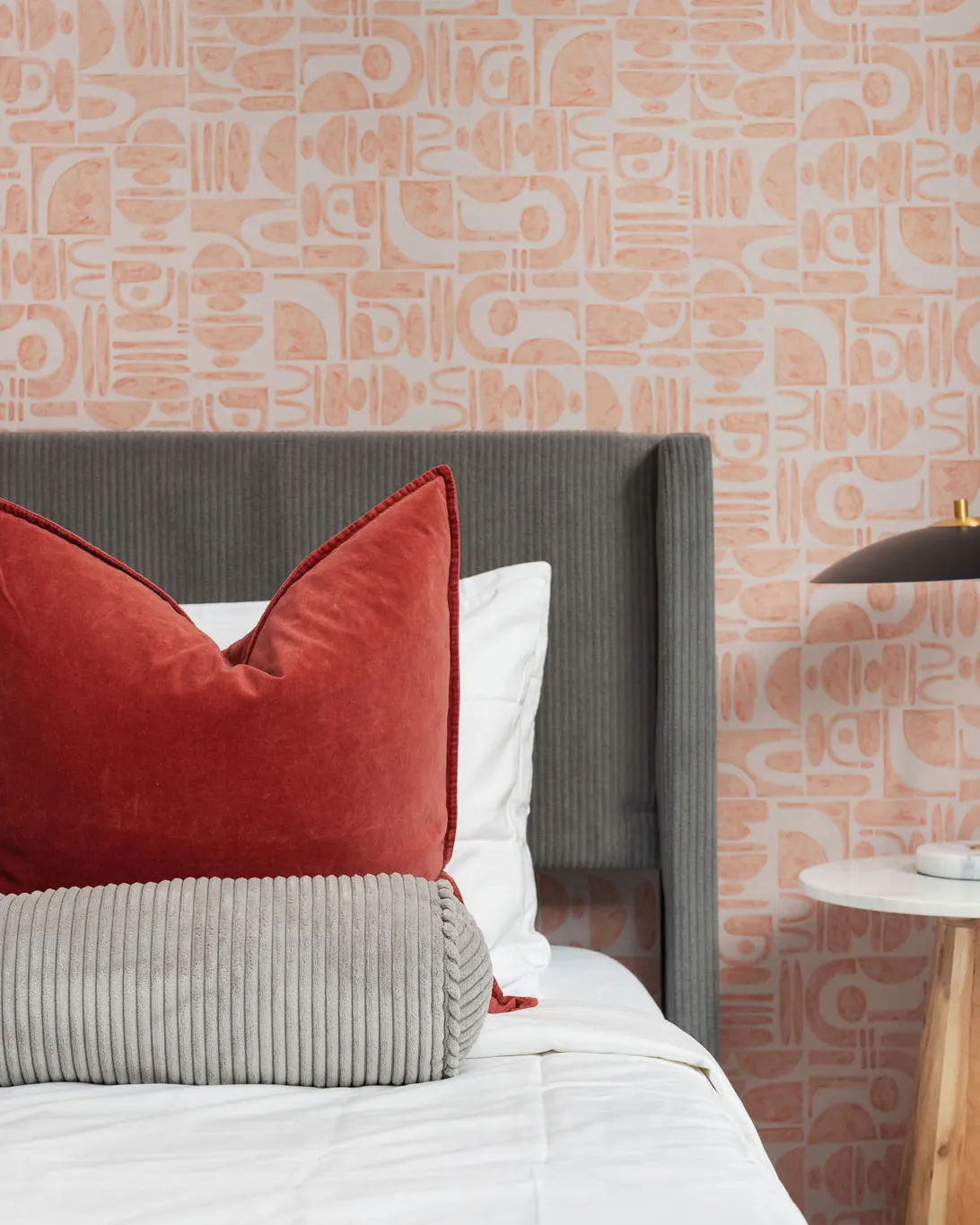 A bed with a grey corduroy headboard and a red velvet pillow against peach wallpaper with an abstract pattern