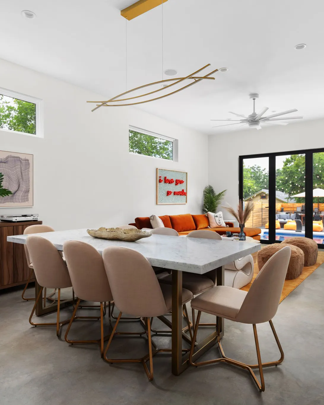 A marble dining table with blush upholstered chairs under a gold pendant, open to the living room and the pool beyond