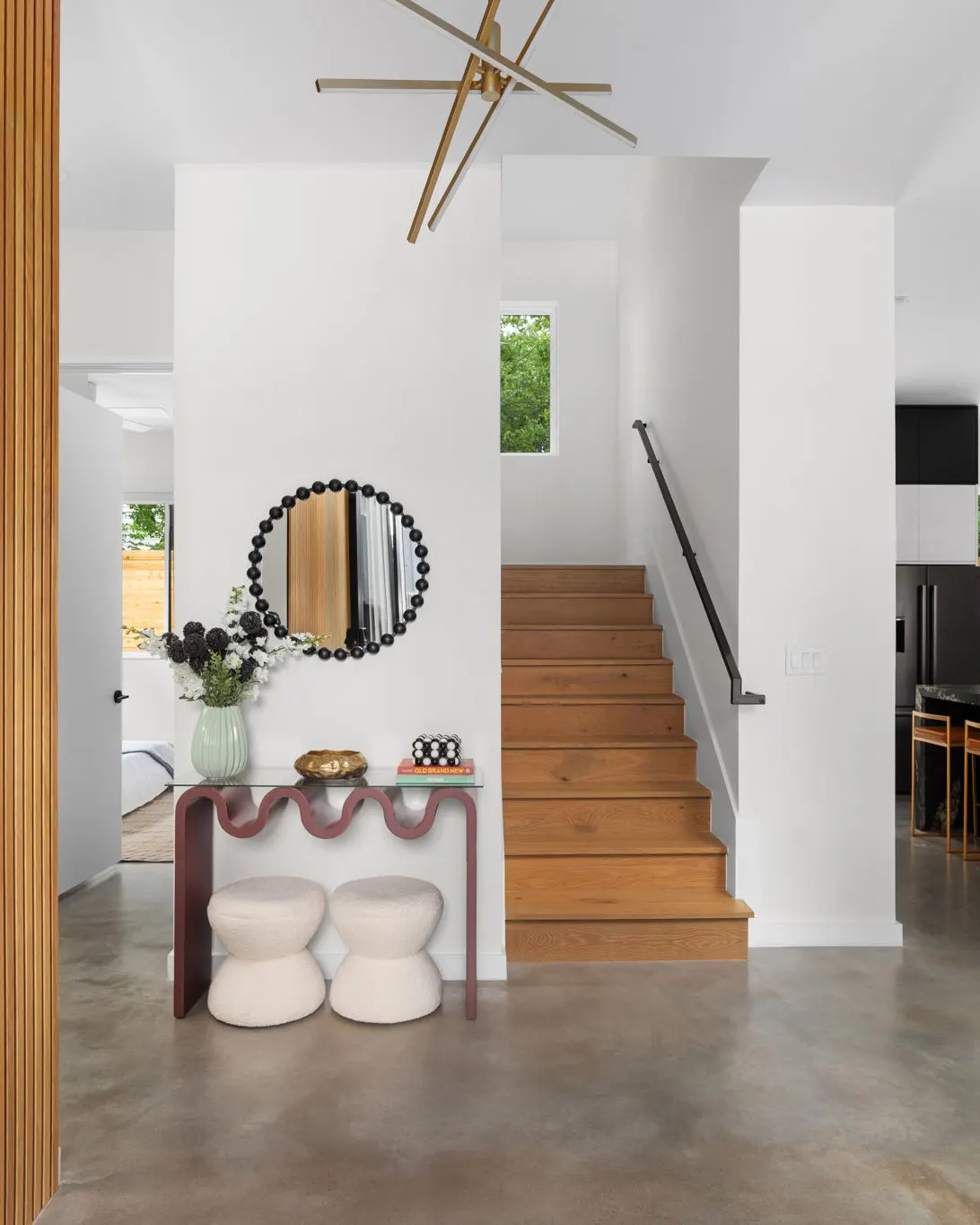 An entry with a wavy burgundy console table, two cream bouclé stools and a round beaded mirror beside an oak staircase