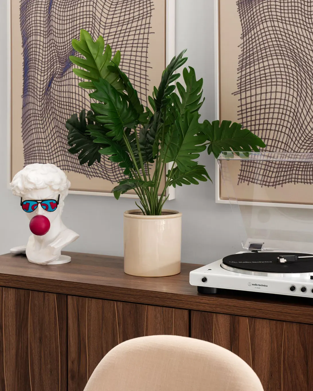 A walnut sideboard with a record player, a potted plant and a classical bust wearing sunglasses and blowing a bubblegum bubble