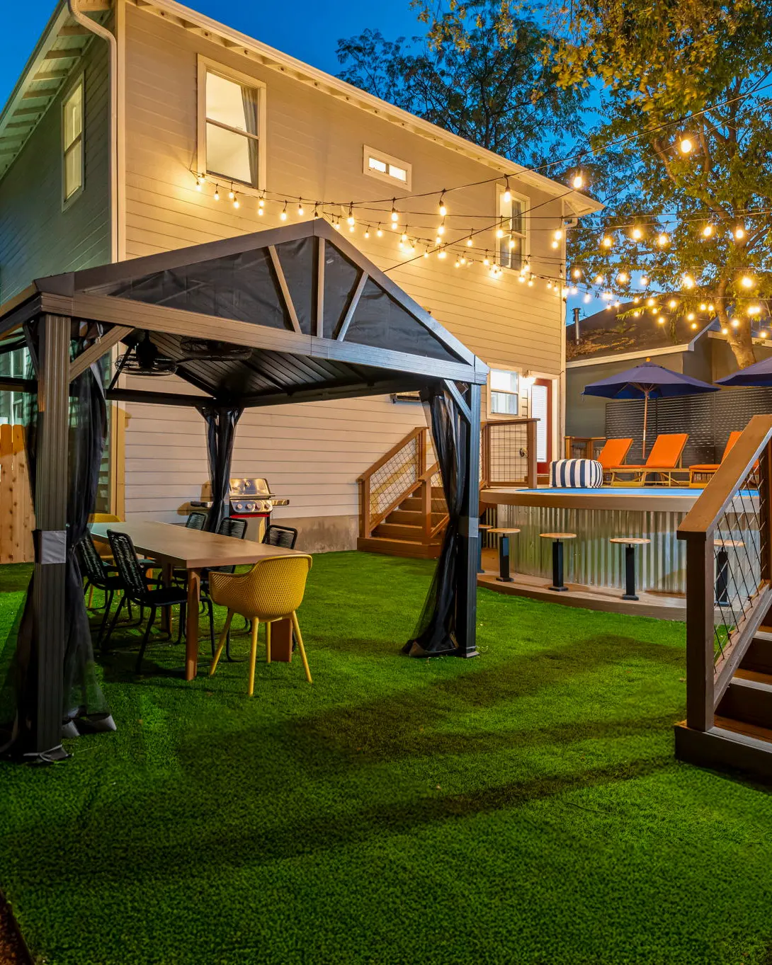 The backyard at dusk under string lights: a pergola over a dining table and grill on the turf, with the pool deck and orange loungers beyond