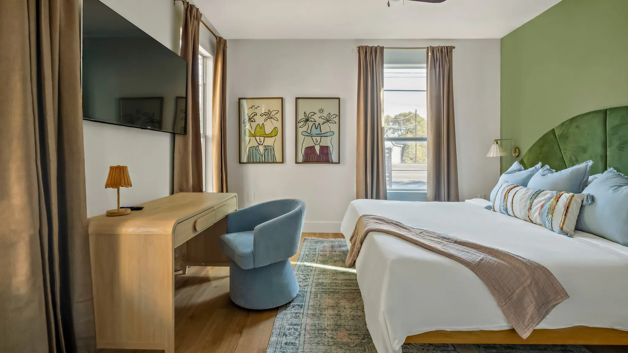 A bedroom with an arched green velvet headboard against a green wall, a curved wooden desk, a blue swivel chair and two prints of women in cowboy hats
