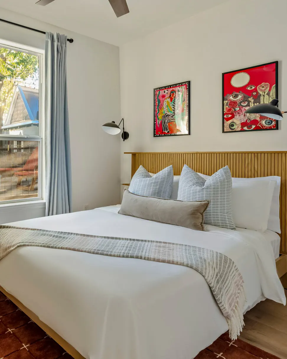 A bedroom with a slatted oak headboard beneath two bold red artworks, one covered in painted eyes