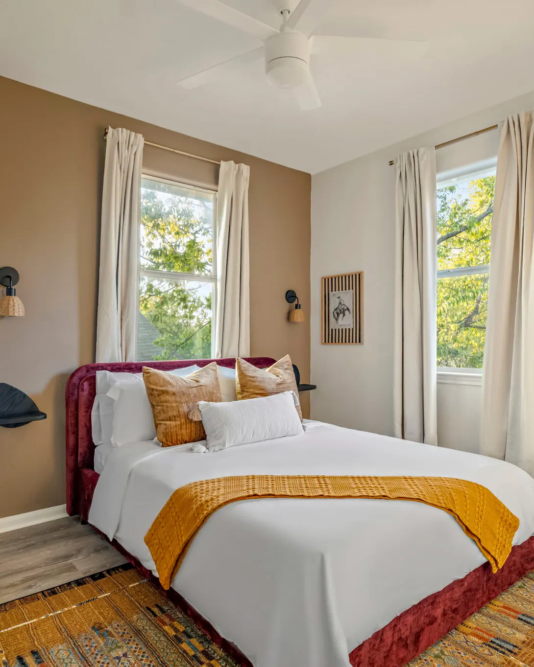 A bedroom with a red velvet bed, a mustard knit throw, a patterned rug and two windows onto the trees