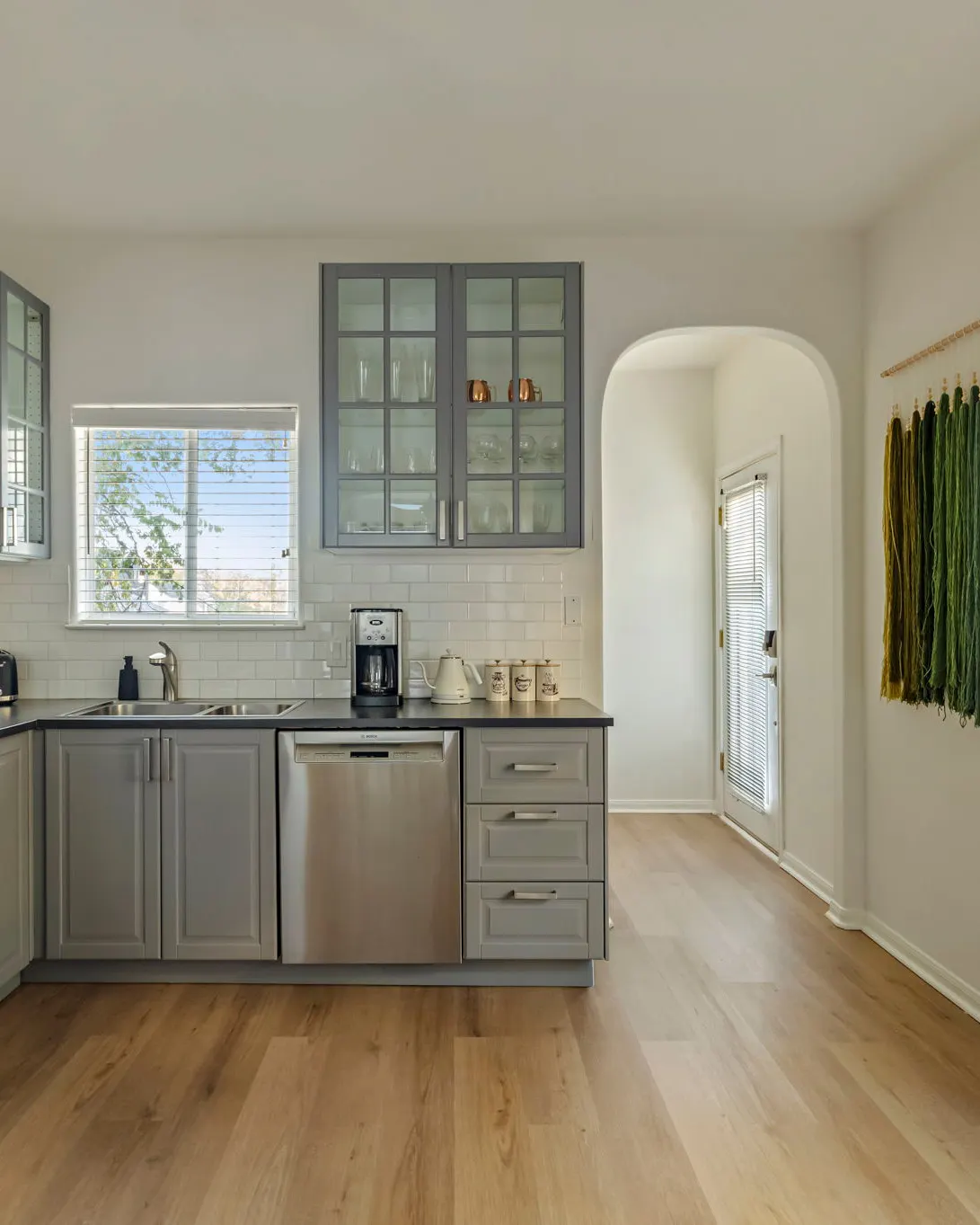A kitchen with grey cabinets, glass-front uppers and a subway tile backsplash, beside an arched doorway and a green yarn wall hanging