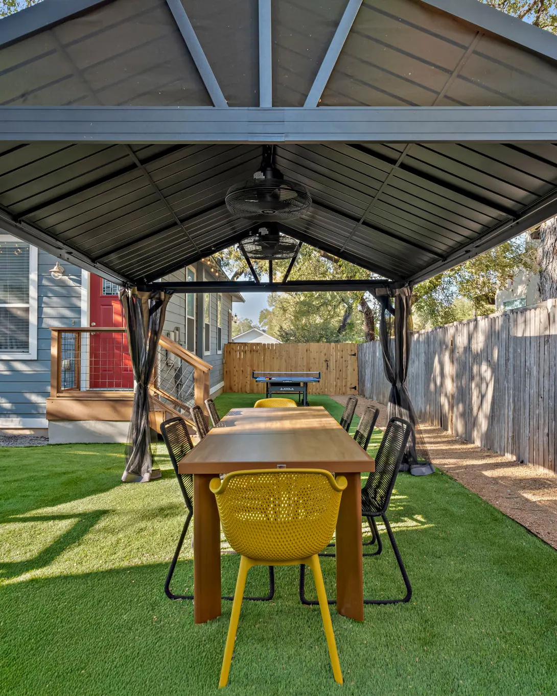 A long dining table with black and yellow chairs under the pergola, with a ping-pong table on the turf beyond
