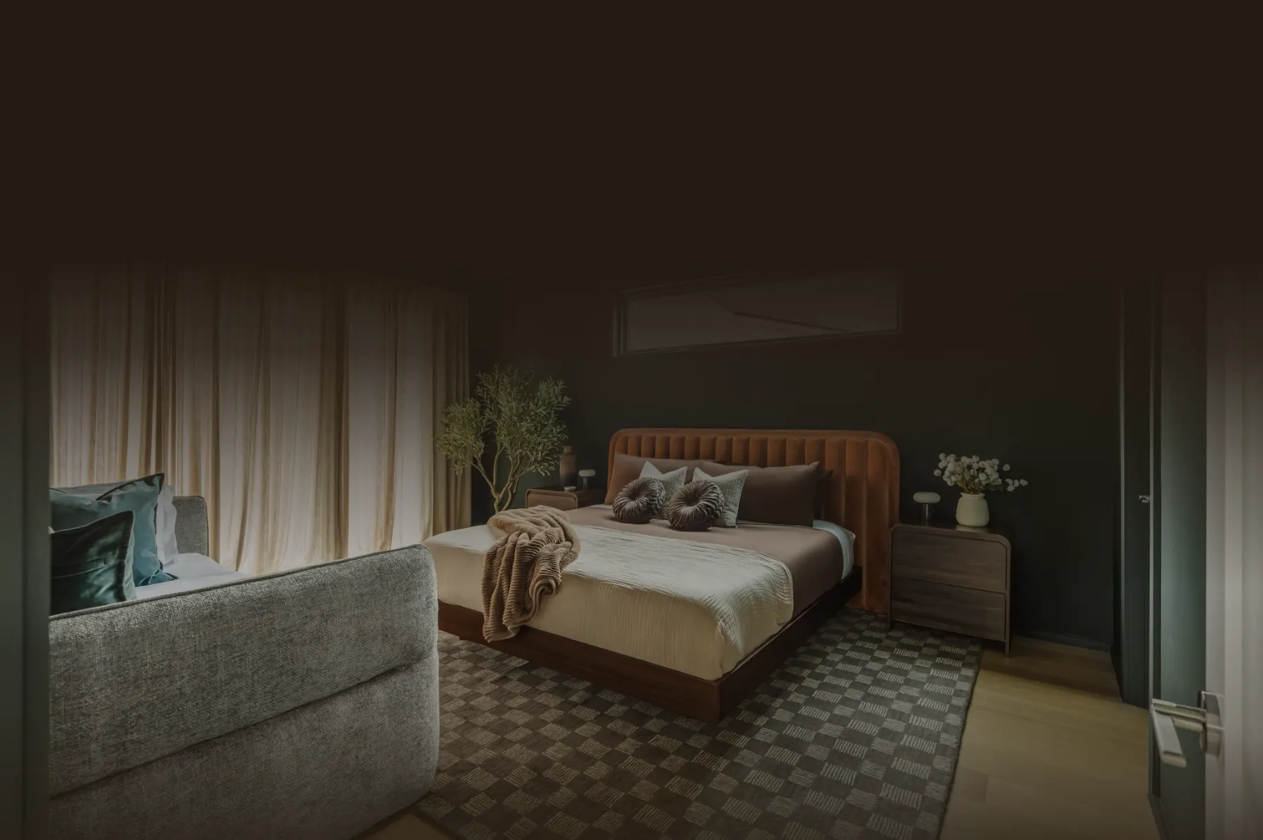 A dimly lit bedroom with a rust velvet headboard, a checked rug and sheer curtains, ready for the next guest