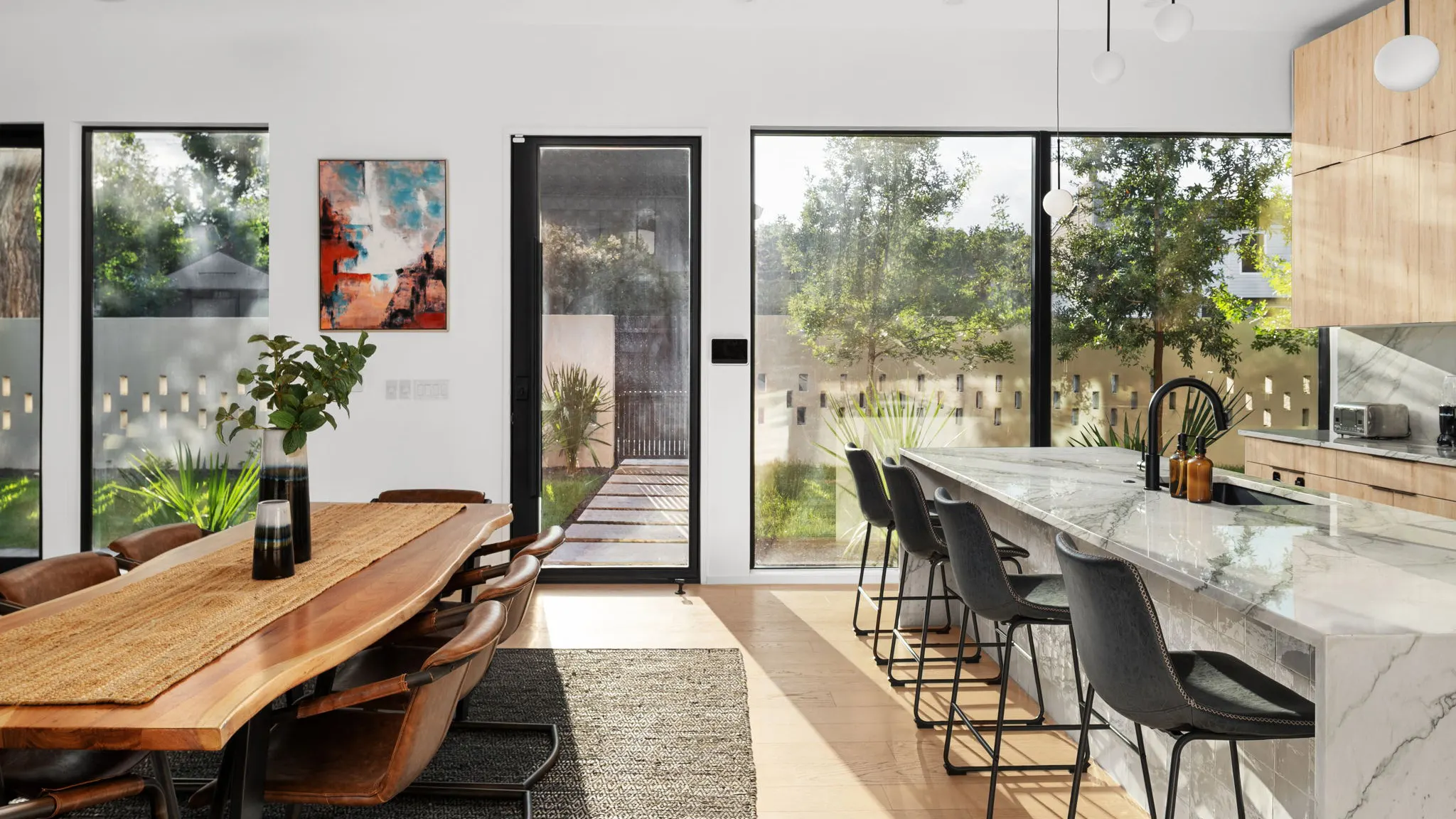 A live-edge dining table with leather chairs beside a marble kitchen island with four bar stools, lit by tall windows onto the garden