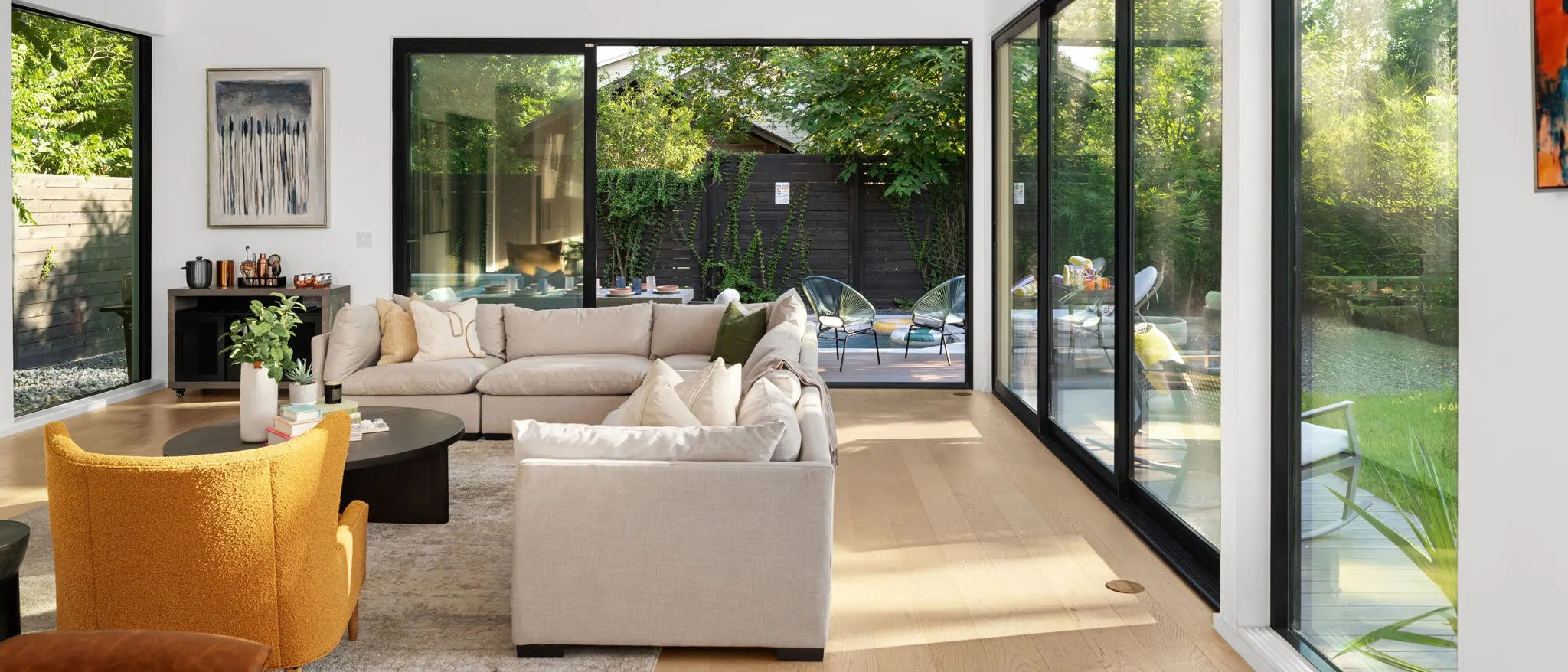 A living room with a cream sectional and a mustard bouclé armchair, walled in glass on two sides, with sliding doors open to the patio and garden