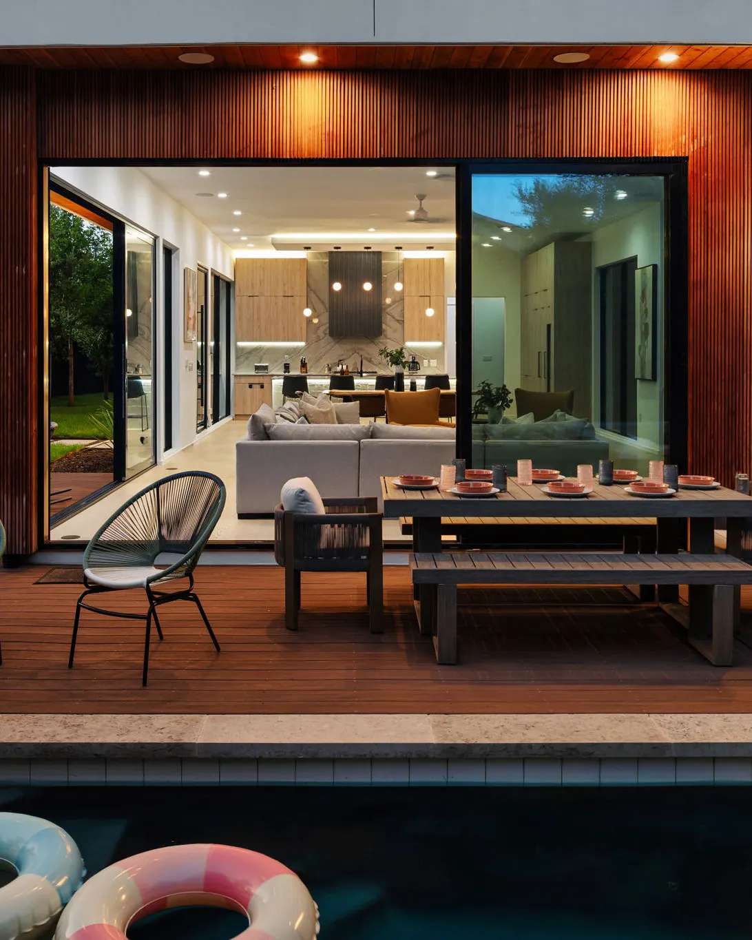 The house at dusk from across the pool: a deck with an outdoor dining table and a rope chair, and wide sliding doors open onto the living room and kitchen