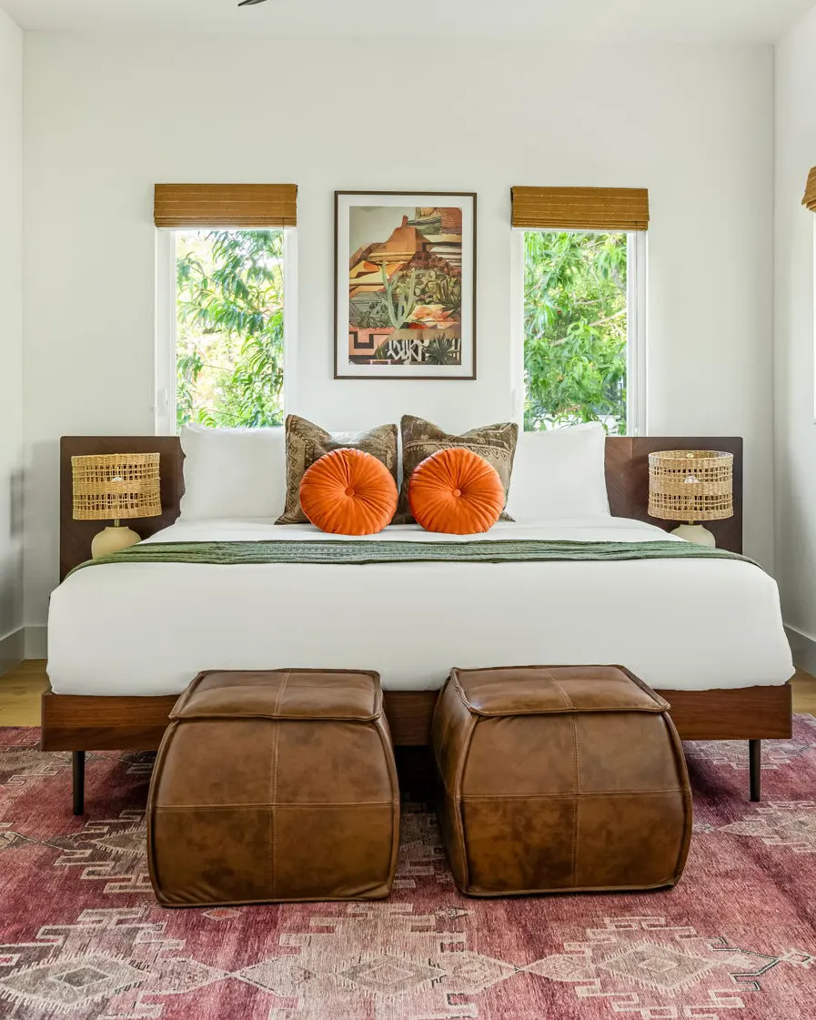 A bed with two round orange velvet pillows between two windows, with leather poufs at its foot on a red patterned rug