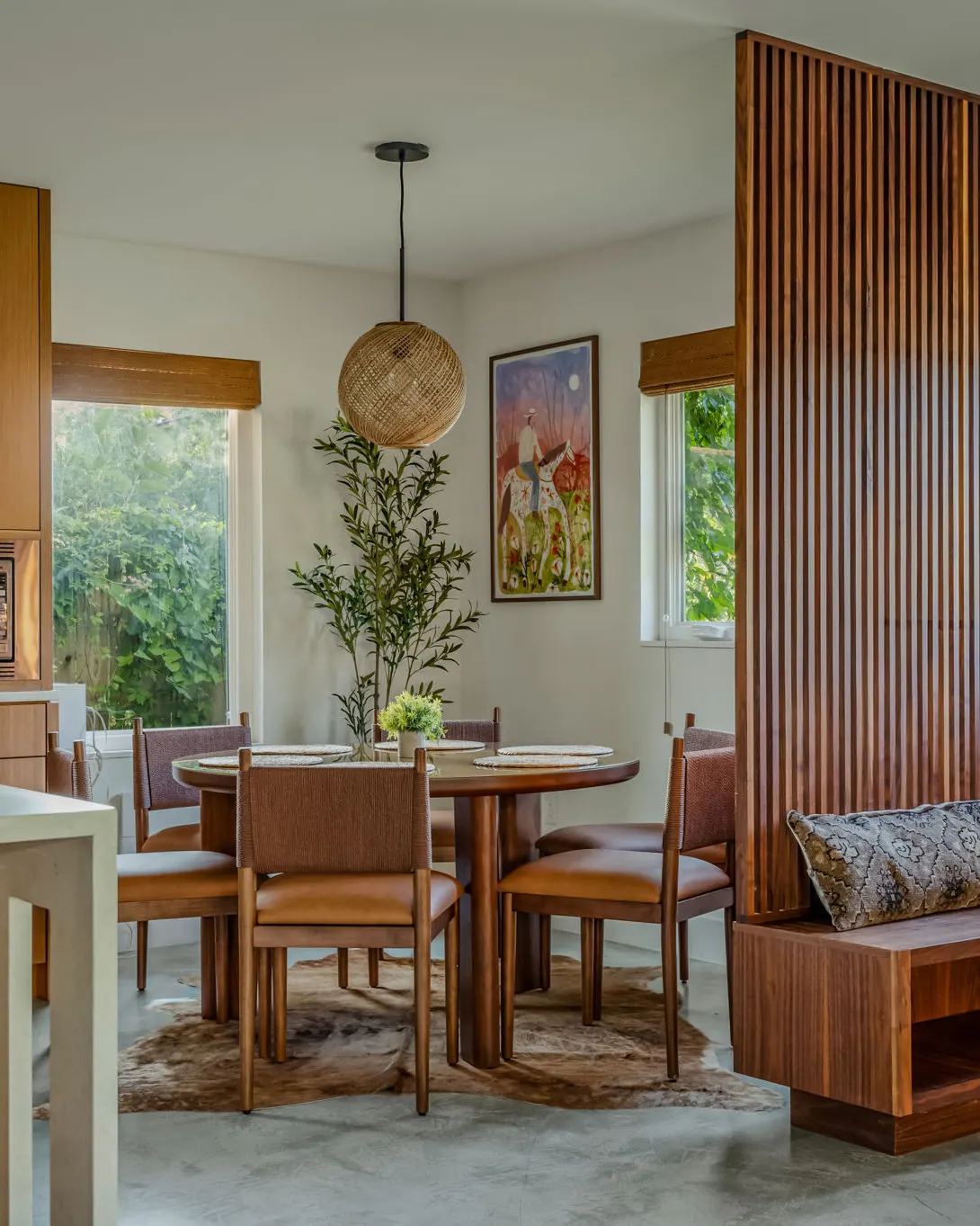 A round dining table with leather chairs under a woven pendant light, on a cowhide rug beside a slatted wood partition and a painting of a rider on horseback