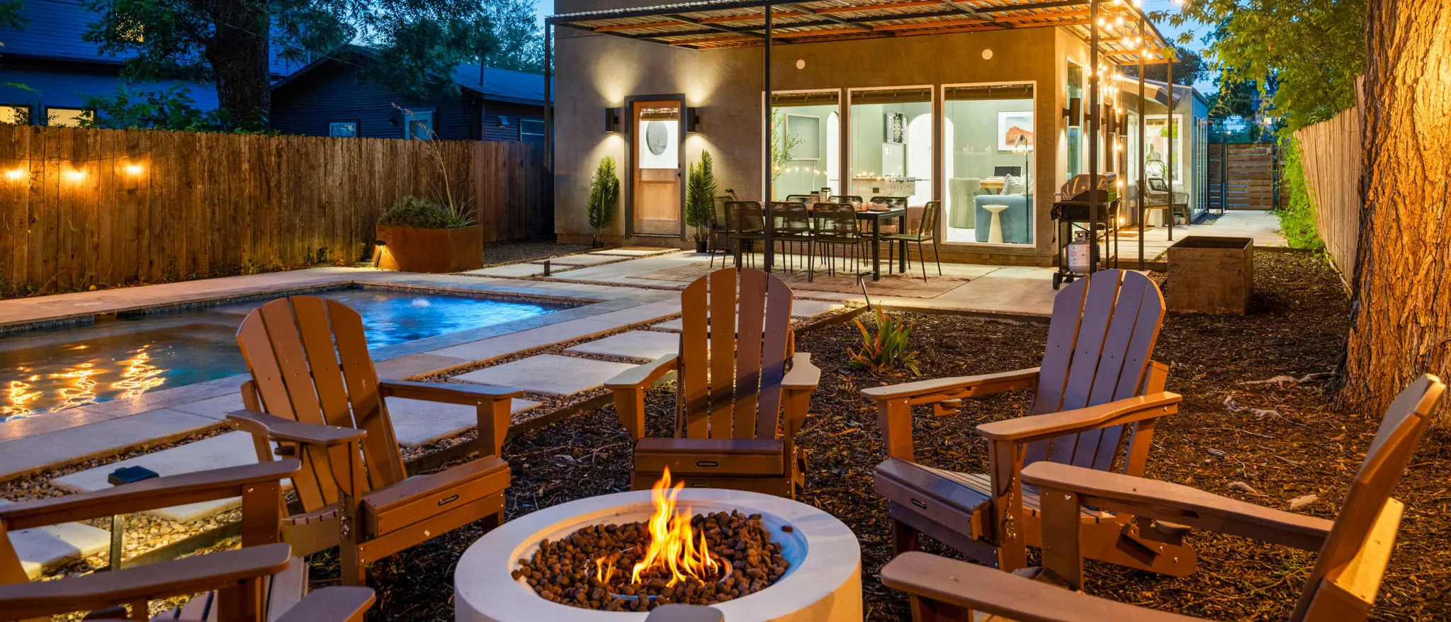 The backyard at dusk: Adirondack chairs around a lit fire pit, the pool, and the house with its dining table under the pergola
