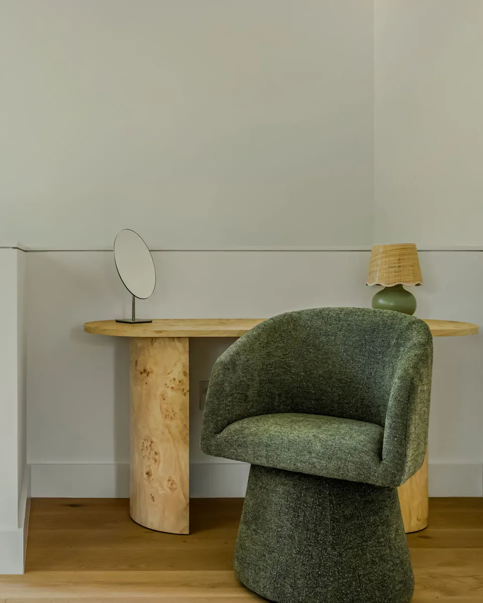 A green bouclé swivel chair at a burl wood desk with a small round mirror and a scalloped lamp