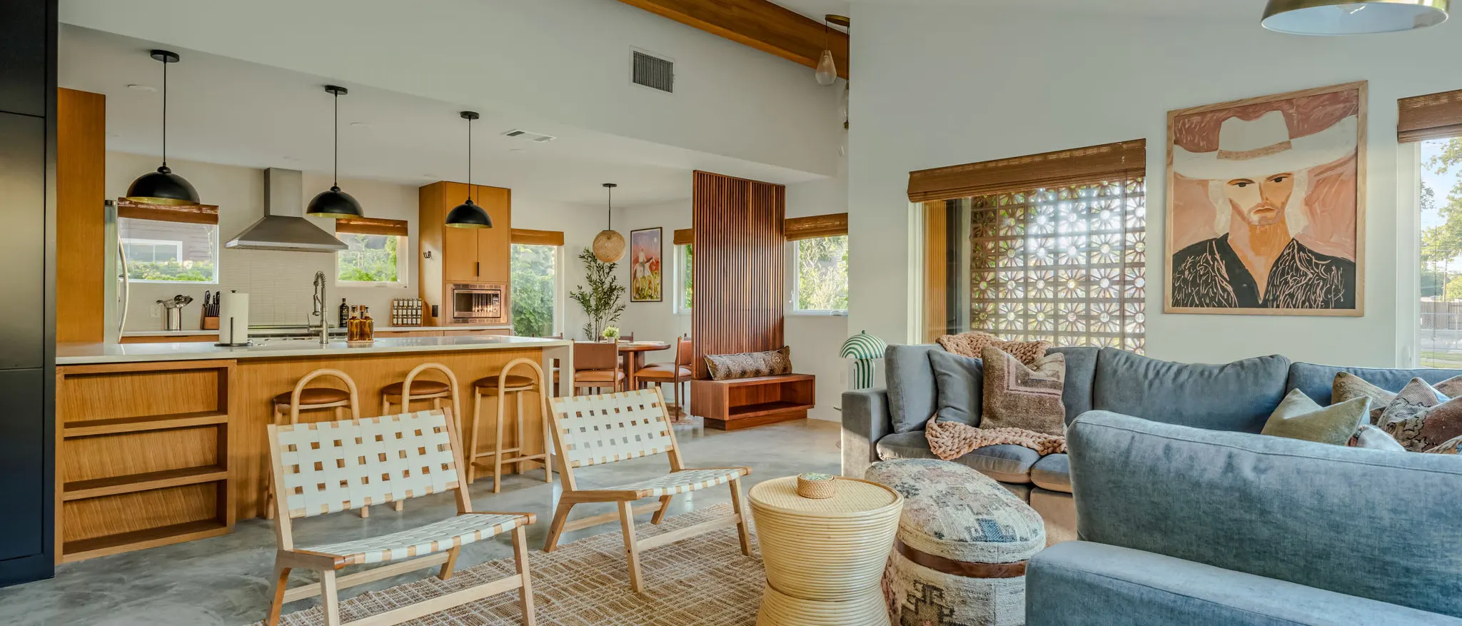 An open-plan kitchen and living room with a warm wood island and woven bar stools, woven lounge chairs, blue sofas, and a large portrait of a cowboy beside a breeze-block window