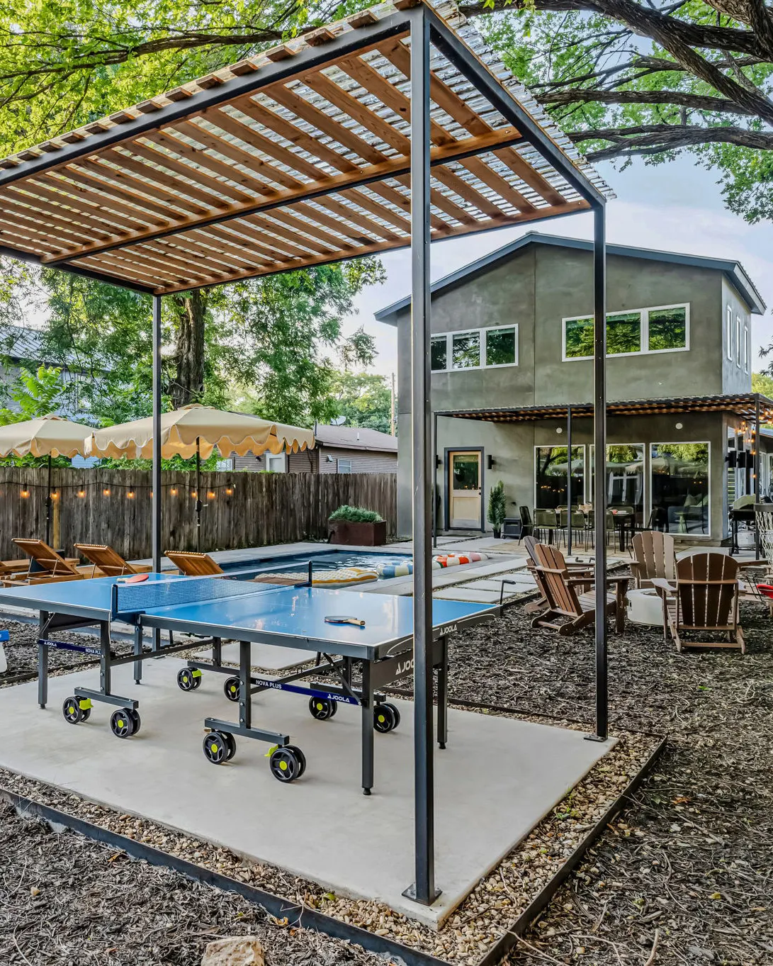 A ping-pong table under a slatted pergola, with Adirondack chairs, the pool and the house beyond