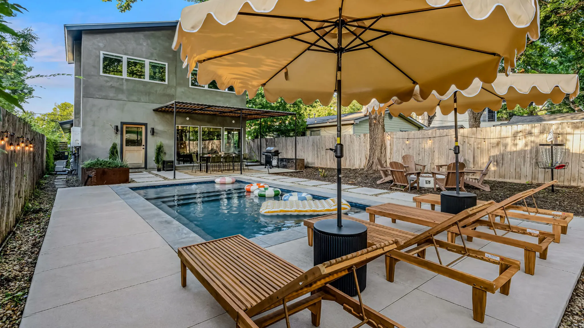The pool with wooden loungers under scalloped umbrellas, Adirondack chairs, a disc golf basket, and a pergola over the dining table by the house