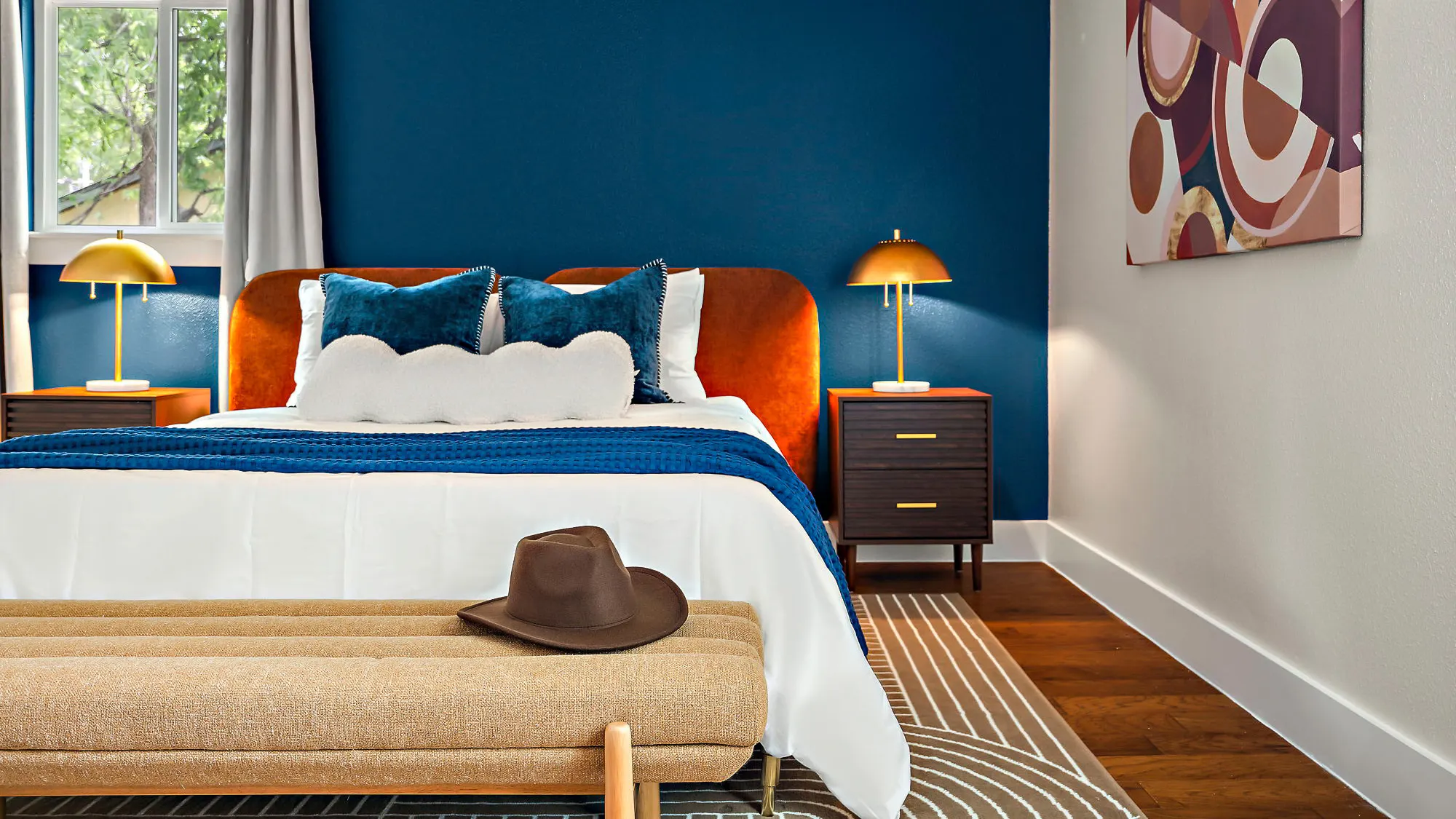 A bedroom with a navy accent wall, a rust velvet headboard, brass lamps, and a cowboy hat on a bench at the foot of the bed