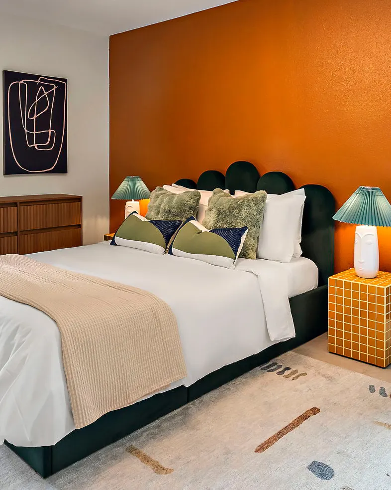 A bedroom with a burnt orange accent wall, a green scalloped velvet headboard, pleated teal lamps and a mustard tiled nightstand