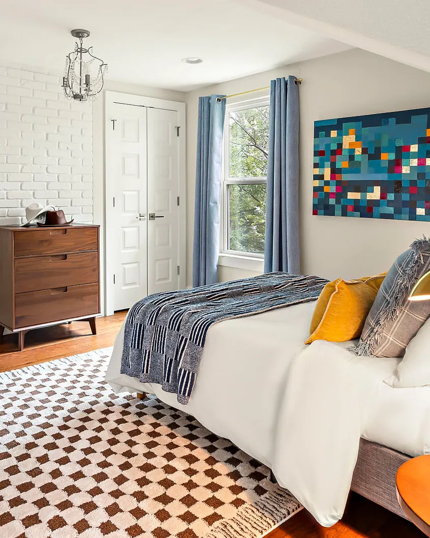 A bedroom with a pixelated blue artwork, blue curtains, a white brick wall and a brown and cream checkered rug