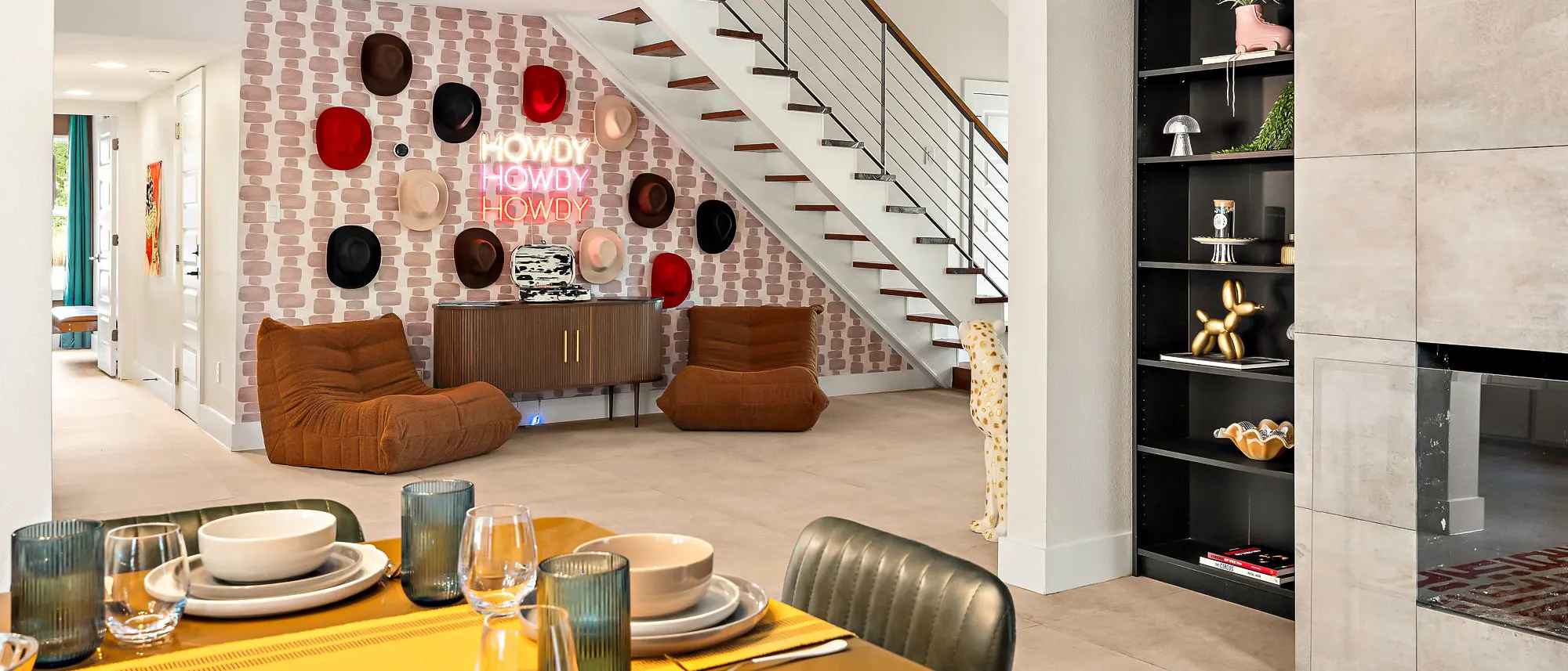 An open-plan living space with a wall of cowboy hats around a neon "Howdy" sign under the staircase, two tan lounge chairs, and a table set with glassware in the foreground