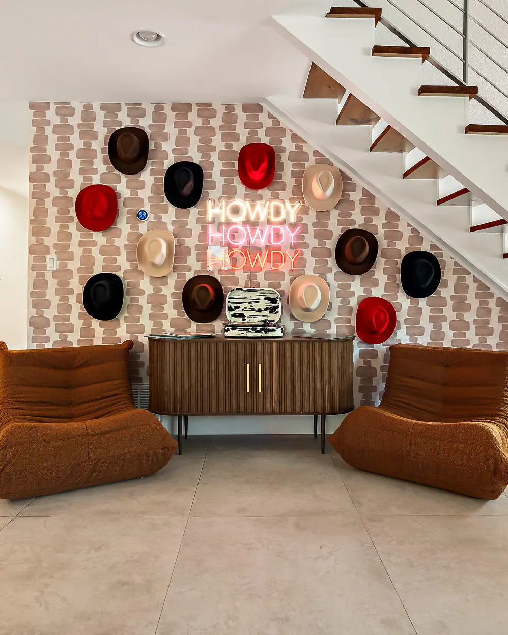 Cowboy hats in red, black, brown and cream arranged around a neon "Howdy" sign, above a fluted sideboard between two tan lounge chairs