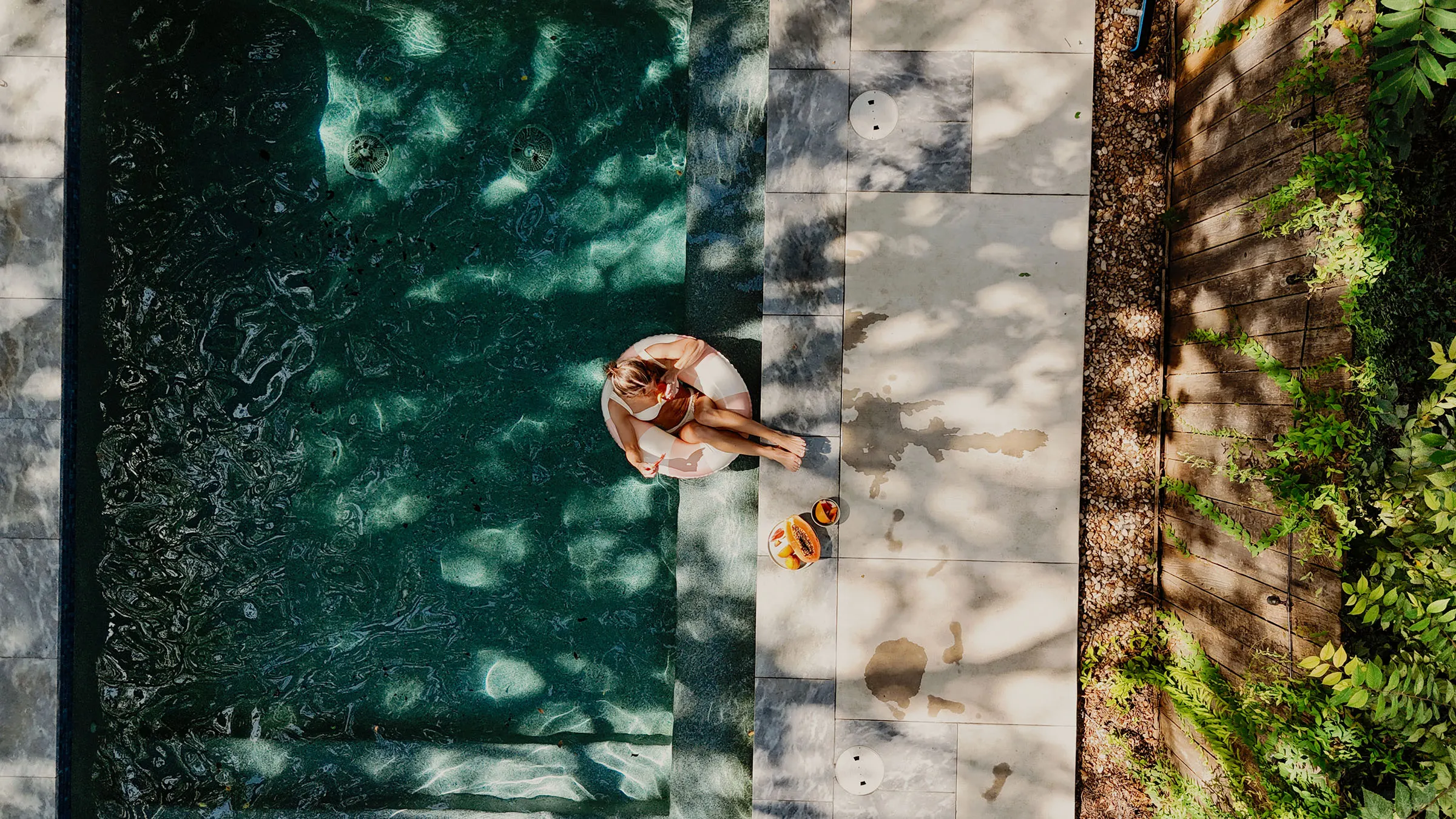 Overhead view of a guest floating on an inflatable in a dark green pool beside a stone deck