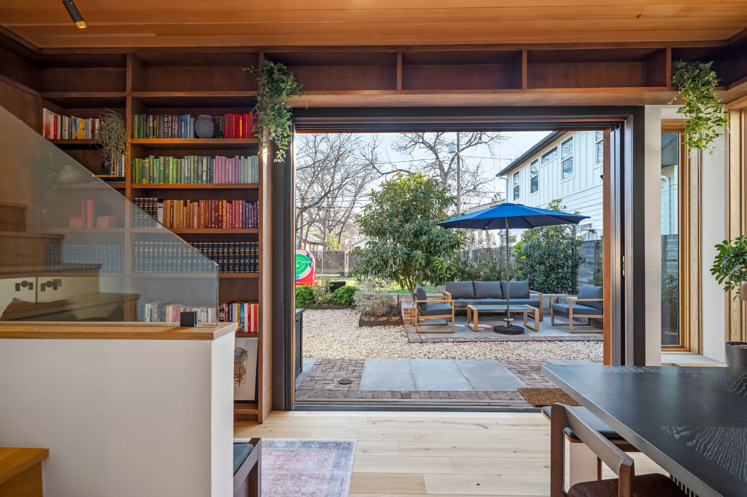 A double-height library with floor-to-ceiling bookshelves, its glass wall folded open onto a gravel patio with a blue umbrella and lounge seating