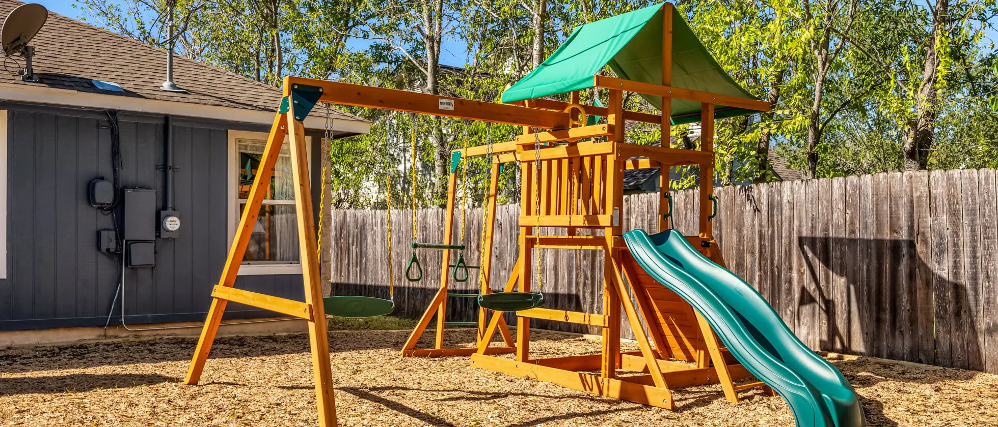 The backyard playground: a wooden play set with swings, a green canopy and a green slide, on wood chips beside the fence