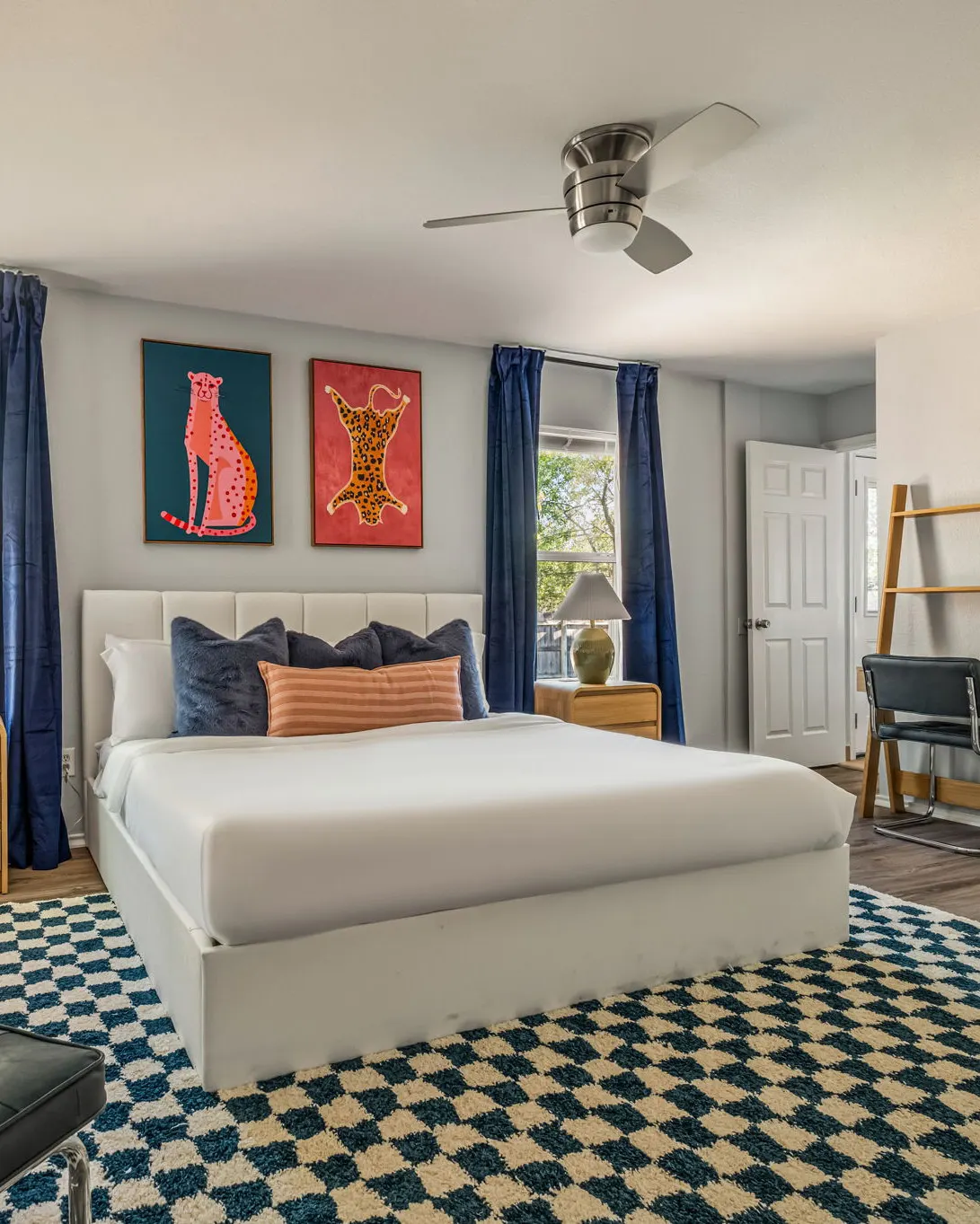 A bedroom with a white upholstered bed, navy pillows and velvet curtains, two leopard prints, and a navy and cream checkered rug
