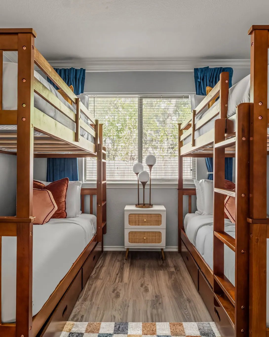 A bunk room with two wooden bunk beds facing each other, blue curtains, a rattan nightstand and a globe lamp beneath the window