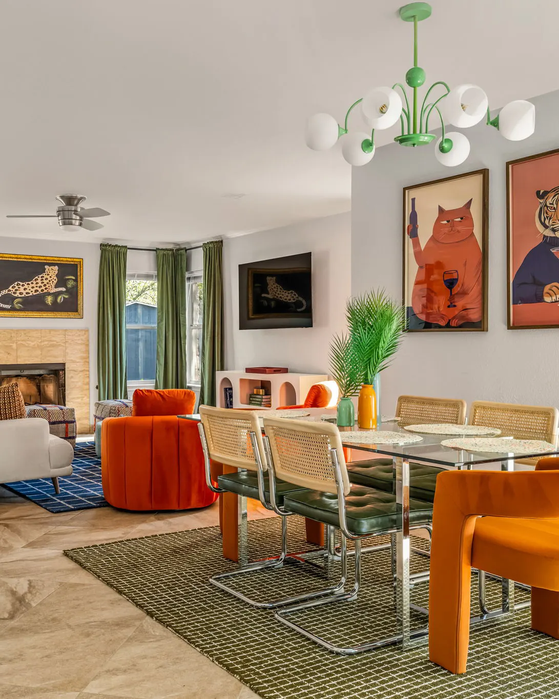 A glass dining table with cane chairs under a green globe chandelier, beneath portraits of a cat drinking wine and a tiger, with orange velvet armchairs and the living room beyond