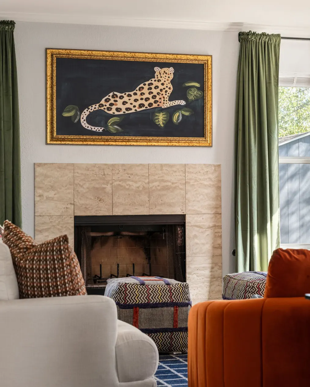 A travertine fireplace beneath a gold-framed painting of a leopard, between green velvet curtains, with a patterned pouf and an orange velvet armchair