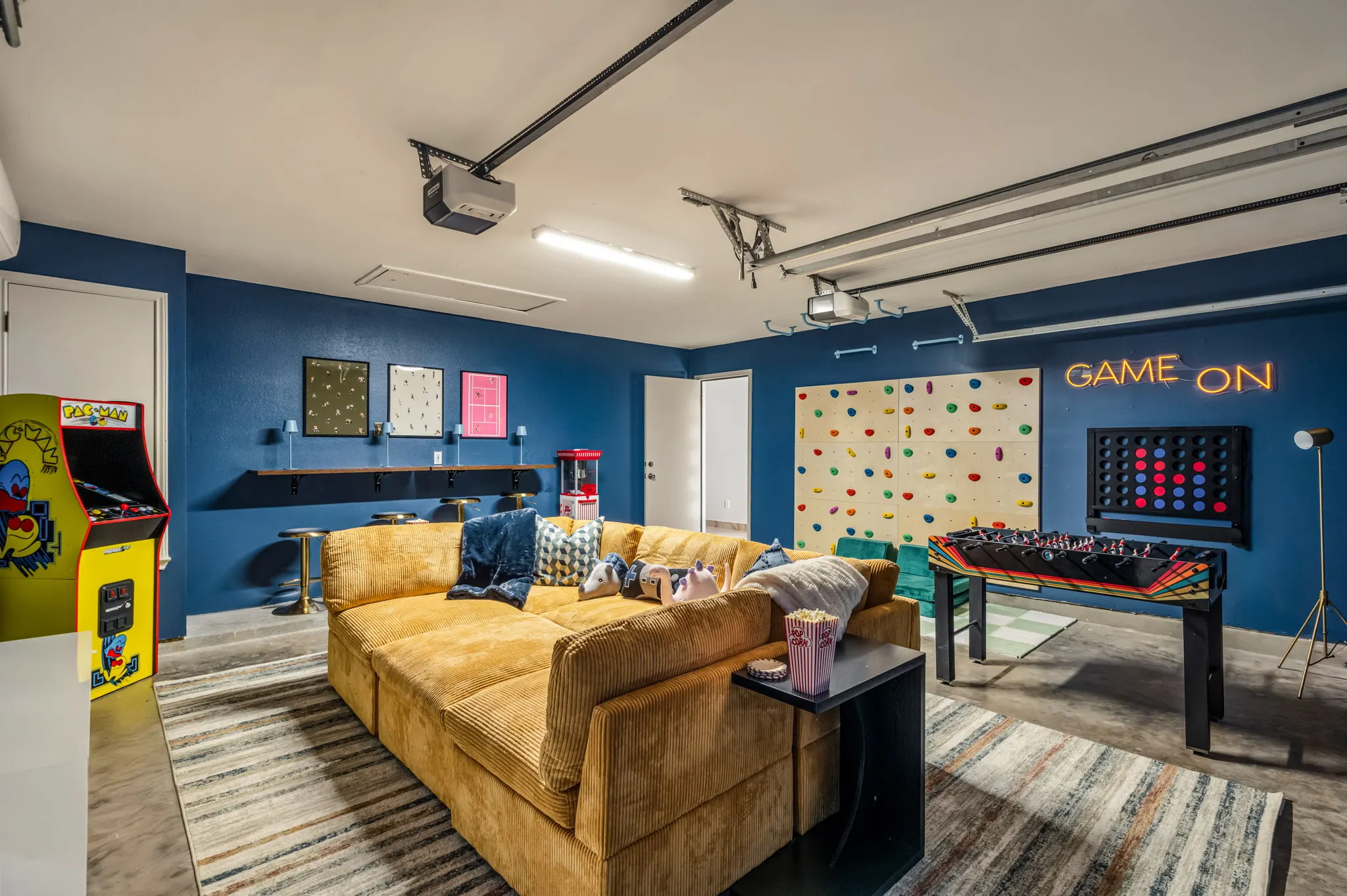 The garage game room: a mustard corduroy sectional facing a climbing wall, with foosball, a giant Connect Four and a neon "Game On" sign on navy walls