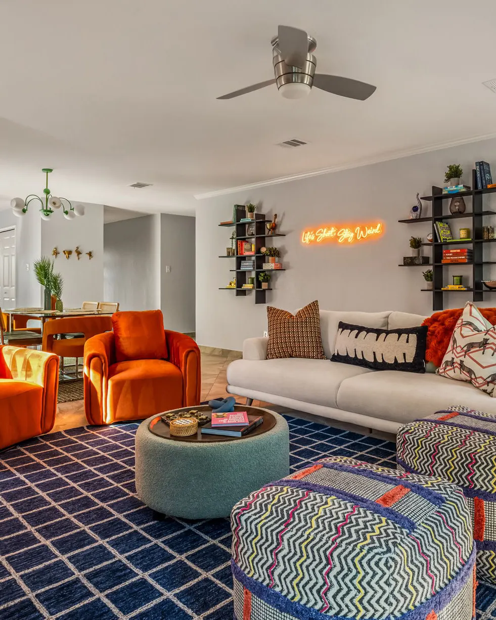 A cream sofa and orange velvet armchairs around a teal ottoman, beneath a neon "Life's Short, Stay Weird" sign between two wall shelves