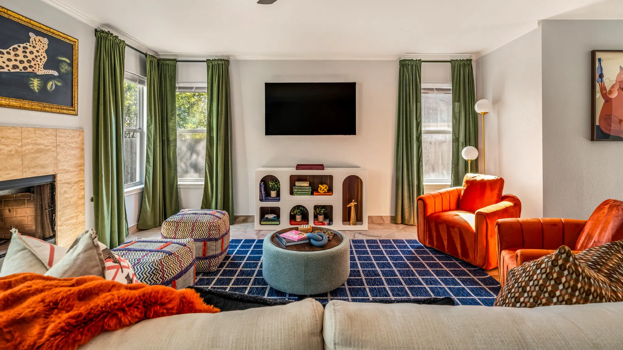 A living room with orange velvet armchairs, zigzag poufs and a round teal ottoman on a navy grid rug, with green velvet curtains and a white arched media unit