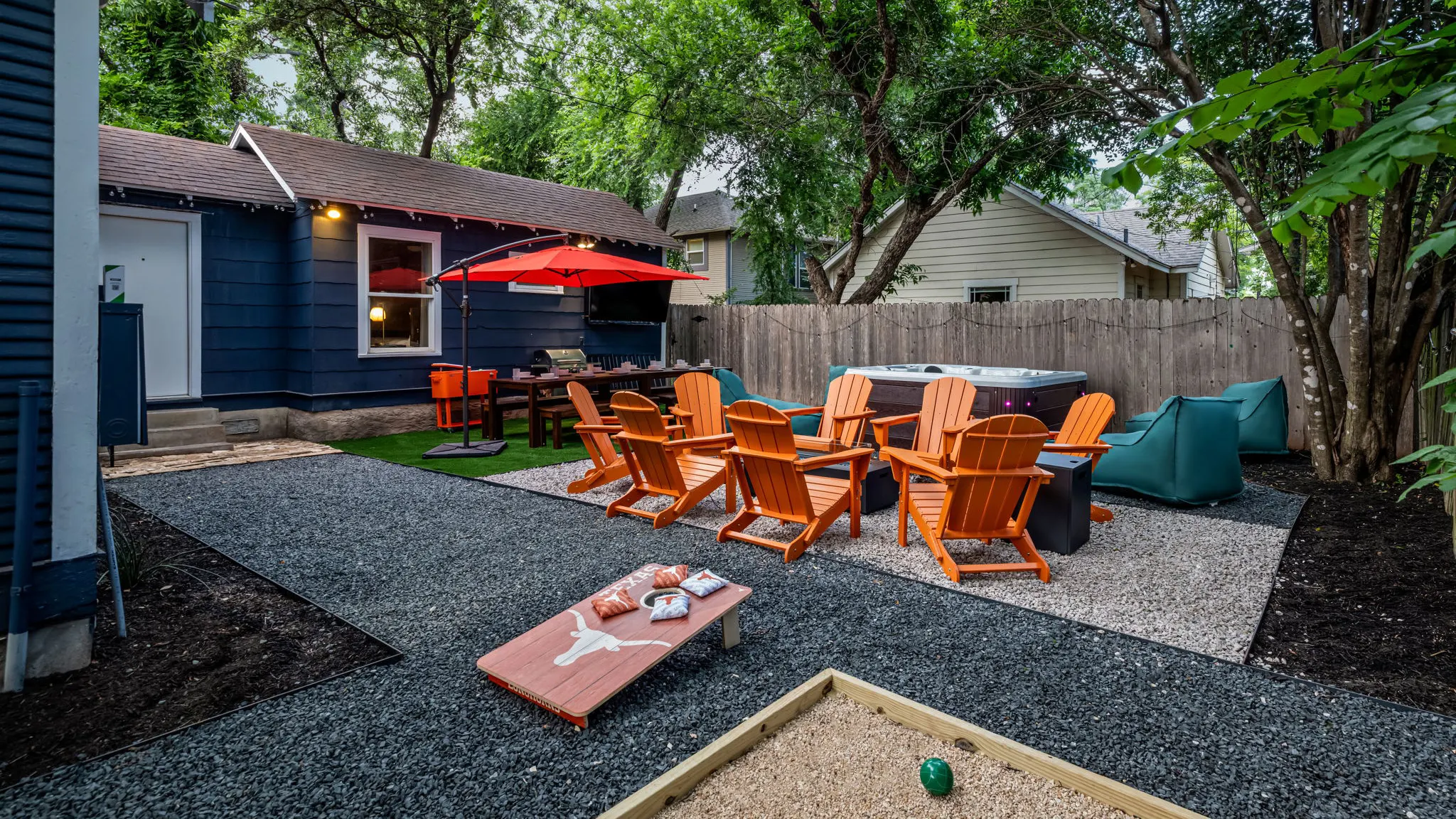 The backyard: orange Adirondack chairs around a fire pit by the hot tub, a Longhorns cornhole board on the gravel, and a dining table under a red umbrella by the house