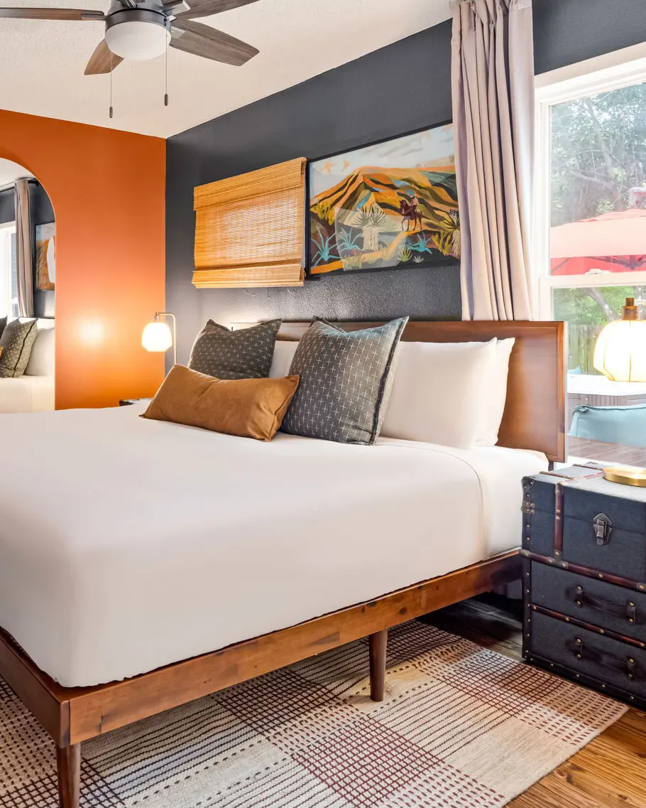 A bedroom with a burnt orange wall and archway, a charcoal accent wall with a desert landscape painting, and a vintage trunk as a nightstand