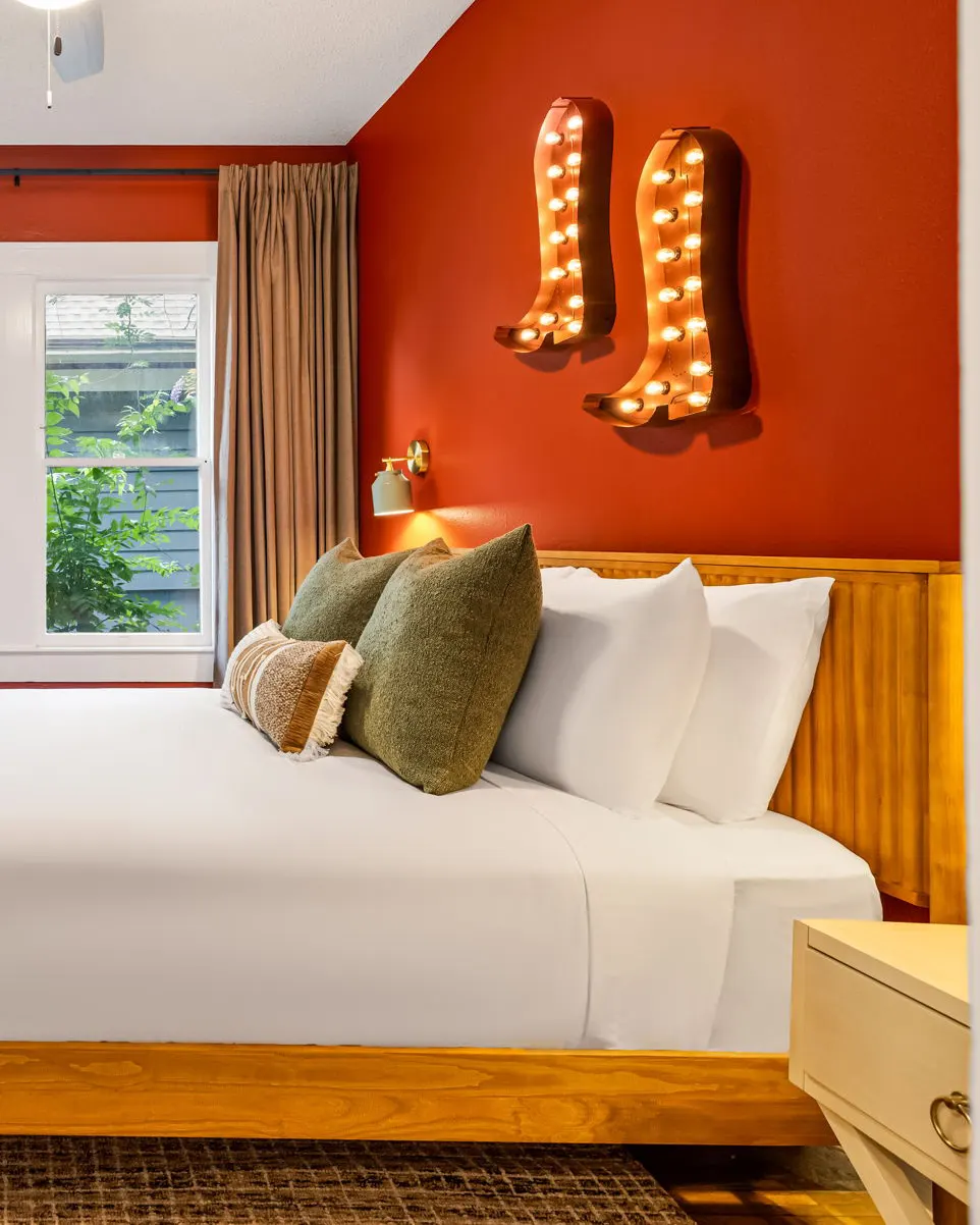 A bedroom with a burnt orange wall and two lit marquee cowboy boots above a wooden headboard