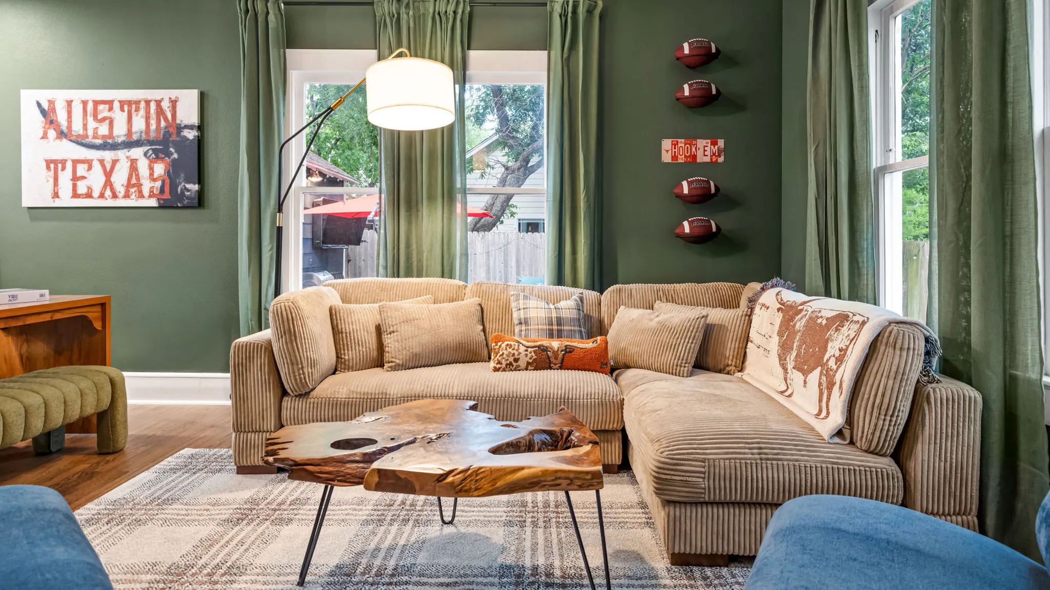 A living room with a corduroy sectional and a live-edge coffee table against a green wall, with footballs mounted around a "Hook 'Em" sign and an "Austin Texas" artwork