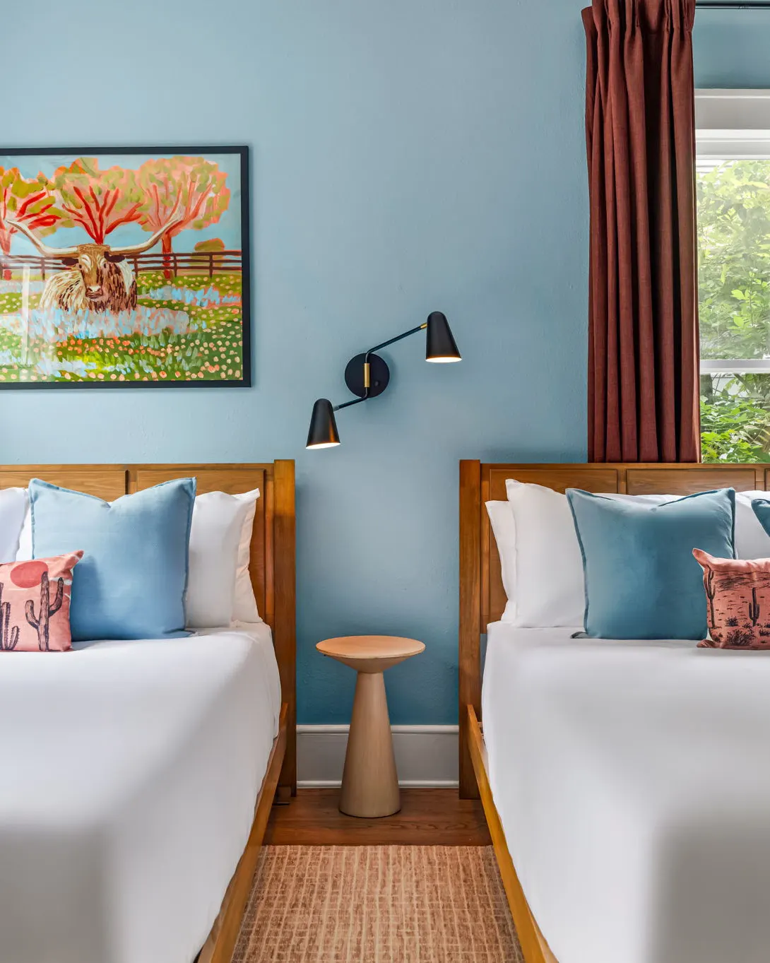 Two beds with blue and cactus-print pillows beneath a colorful painting of a longhorn, against a pale blue wall