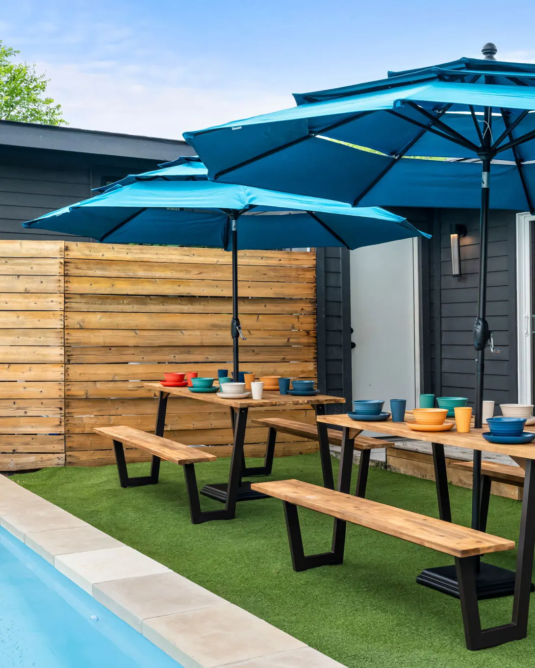 Two picnic tables set with colorful dishes under blue umbrellas, on the turf beside the pool
