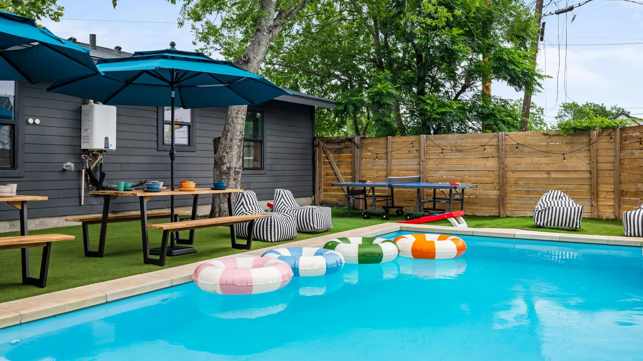 The pool with a row of striped inflatable rings, picnic tables under blue umbrellas, striped bean bag chairs and a ping-pong table on the turf