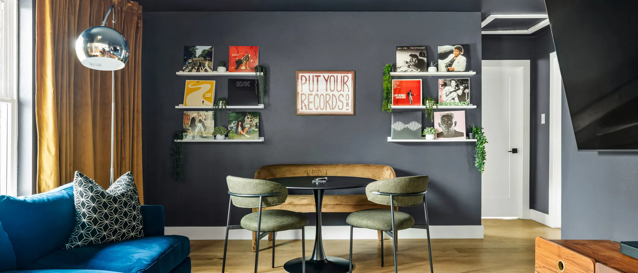 A charcoal living room with album covers on floating shelves around a "Put Your Records On" sign, a bistro table with green chairs, and a blue velvet sofa