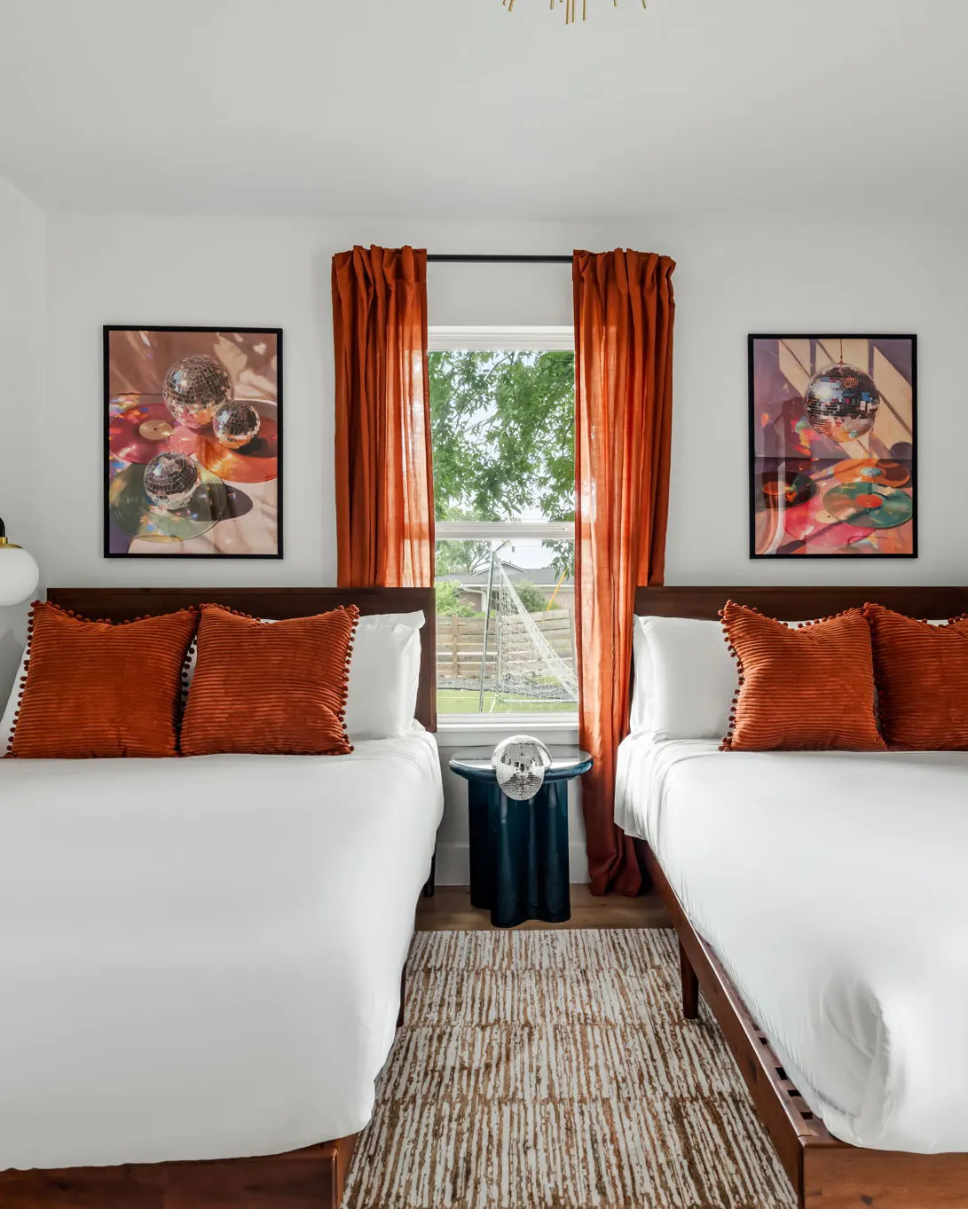 Two beds with rust corduroy pillows beneath photographs of disco balls on vinyl records, on either side of a window with rust curtains