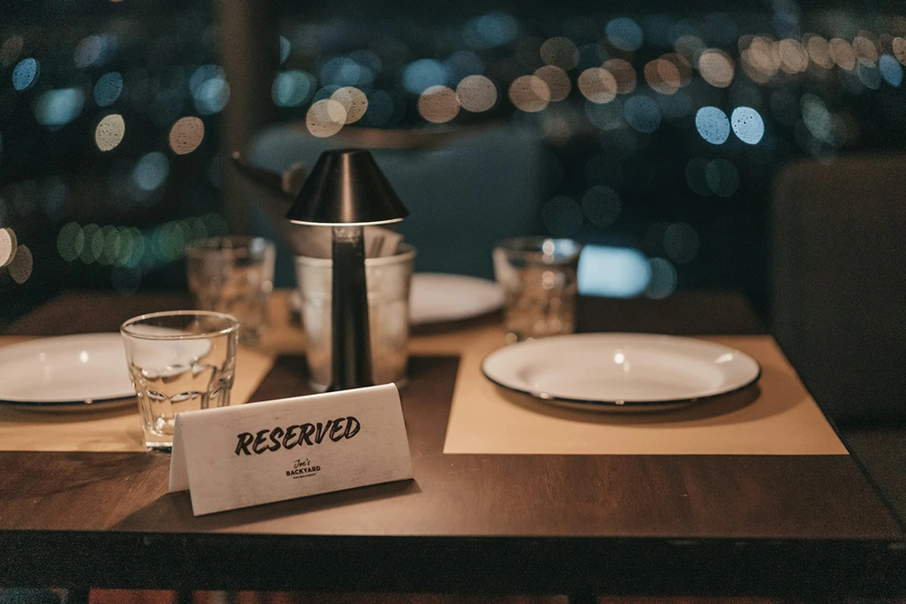 An intimate restaurant table setting with a 'Reserved' sign, two plates, glasses, and a candle holder, with blurred city lights in the background.