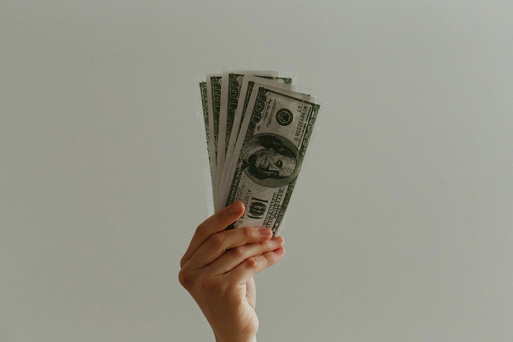 A hand holding a fan of one hundred dollar bills against a neutral background.